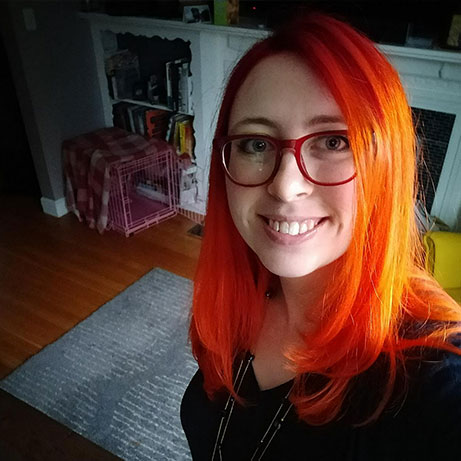 A white woman with with red glasses and long, bright red-orange hair, smiling in her living room.